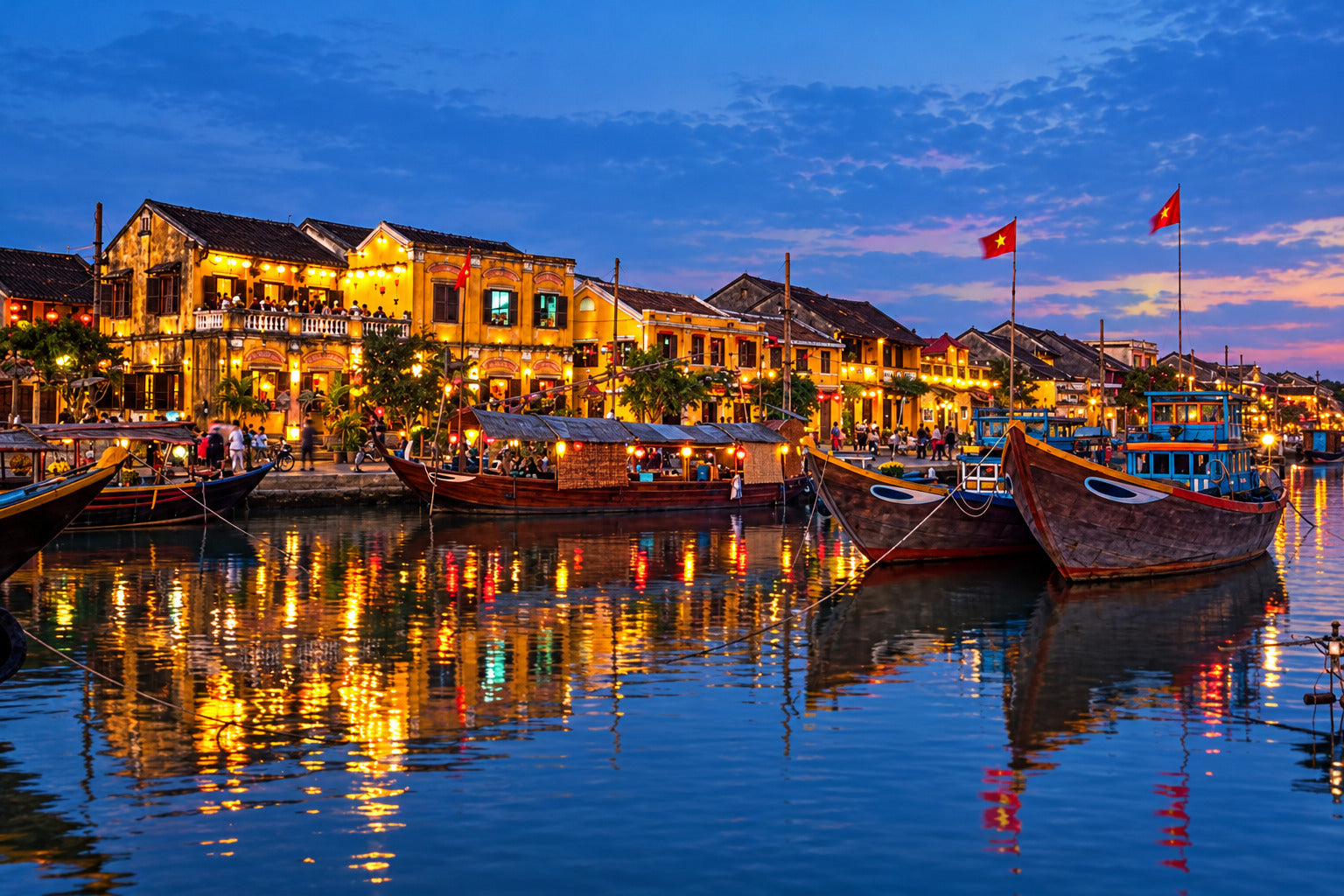 A scenic waterfront at dusk with traditional buildings lit in warm yellow, wooden boats moored in the harbor, and reflections on the water. Two red flags are visible above the boats.
